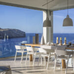 Mesa de comedor con sillas blancas, lámparas colgantes y esquina acristalada abierta al mar y la costa rocosa