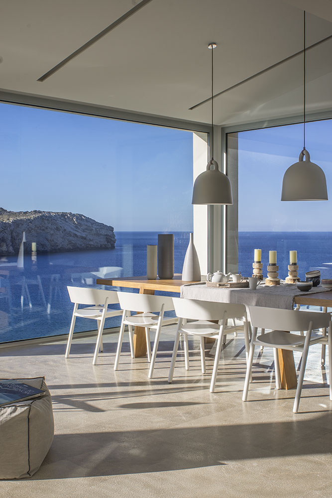 Mesa de comedor con sillas blancas, lámparas colgantes y esquina acristalada abierta al mar y la costa rocosa