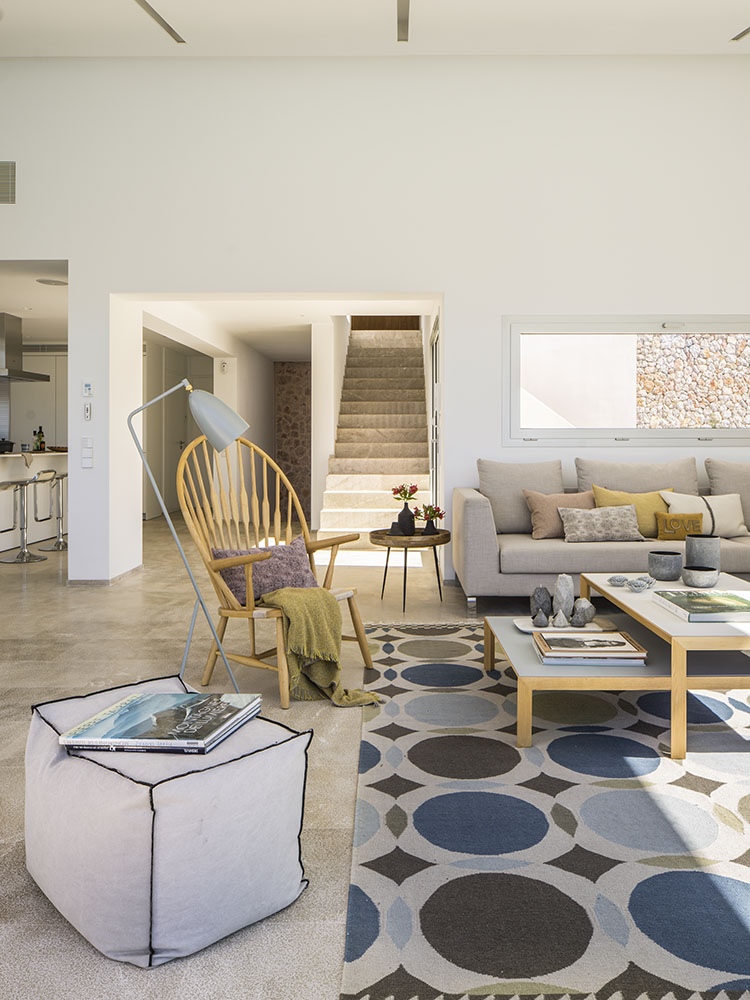 Salón con escalera de piedra al fondo, butaca de madera, puf, sofá beige y alfombra de círculos azules
