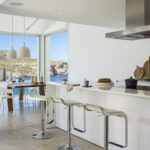 Isla de cocina blanca con taburetes cromados, campana de acero y ventanal al fondo con vistas al mar