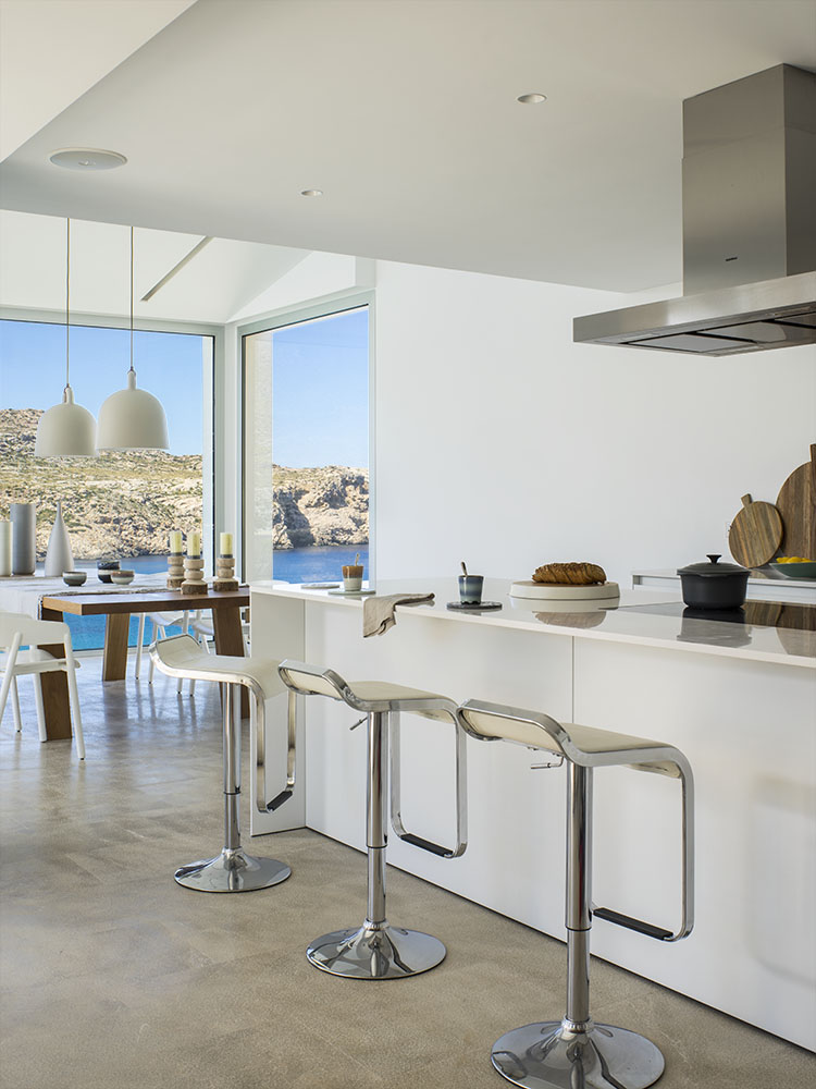 Isla de cocina blanca con taburetes cromados, campana de acero y ventanal al fondo con vistas al mar