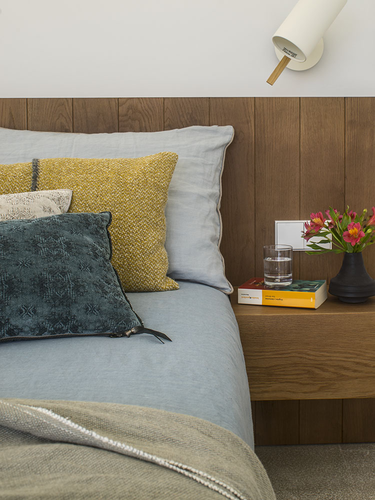 Detalle de la cama con cabecero de madera, cojines mostaza y verde azulado, mesita flotante y flores