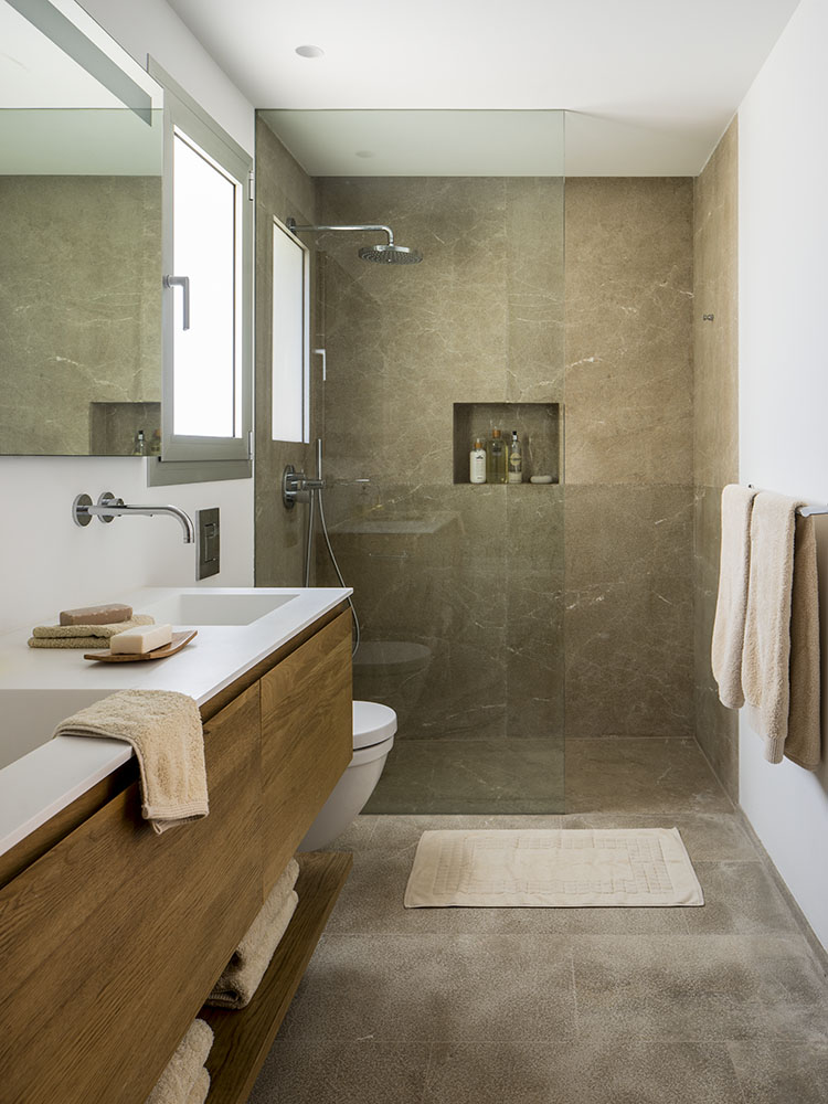 Baño con ducha a ras de suelo revestida en piedra beige, mueble de madera y lavabo blanco