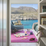 Habitación infantil con alfombra rosa, juguetes, butaca azul y cama, con vistas a la cala y la montaña