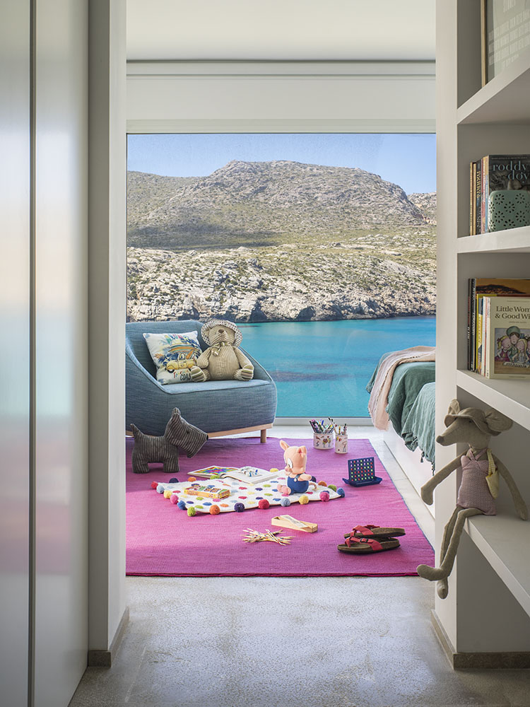 Habitación infantil con alfombra rosa, juguetes, butaca azul y cama, con vistas a la cala y la montaña