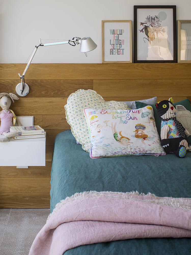Cama infantil con cabecero de madera, ropa de cama verde, colcha rosa, cojines estampados y lámpara articulada
