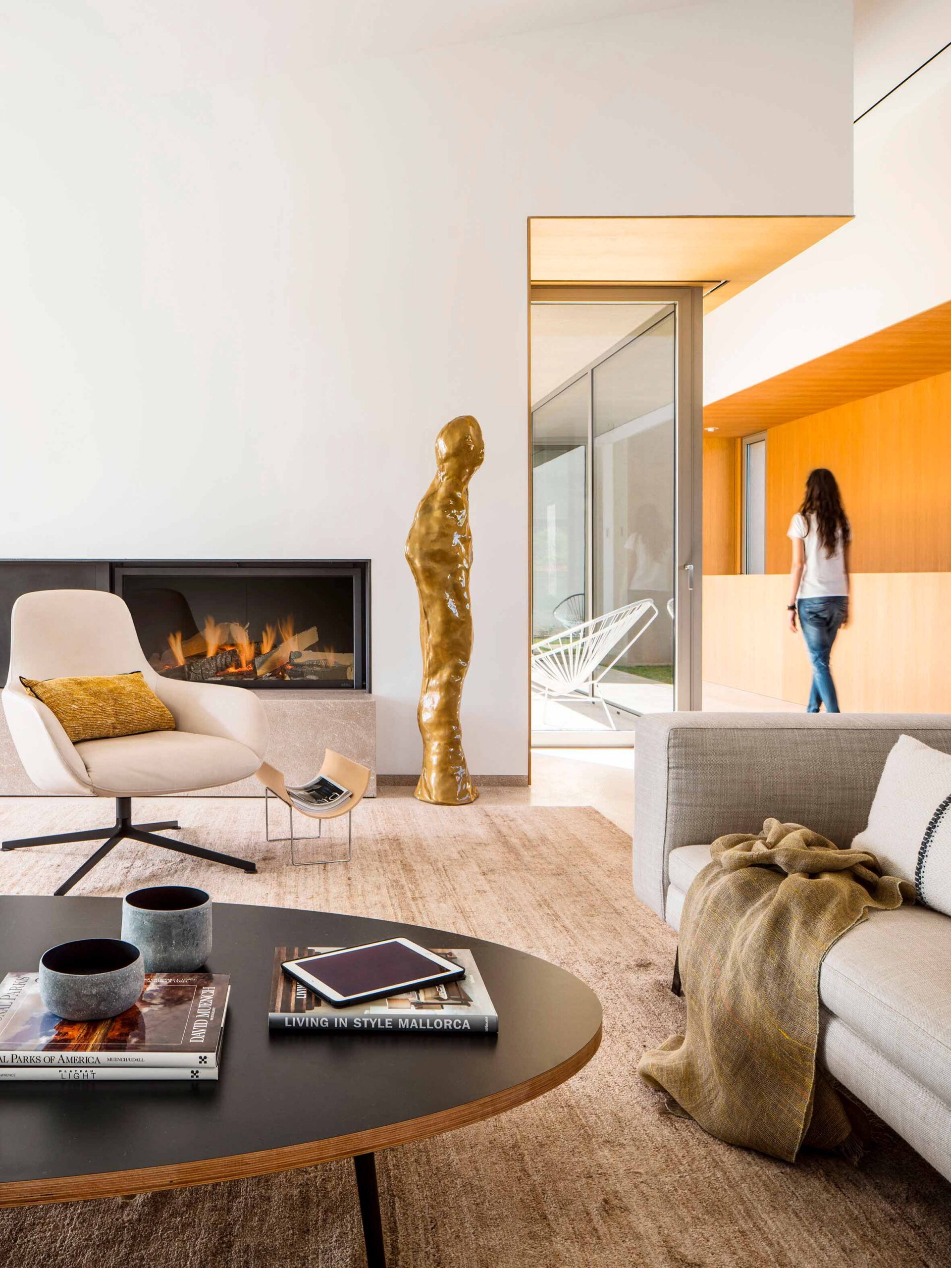 Salón con chimenea empotrada, sillón crema, escultura dorada y paredes de madera al fondo con puerta acristalada