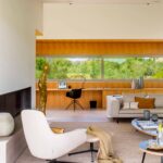 Rincón de despacho en madera clara con ventanal a la vegetación, sofá claro y sillón blanco