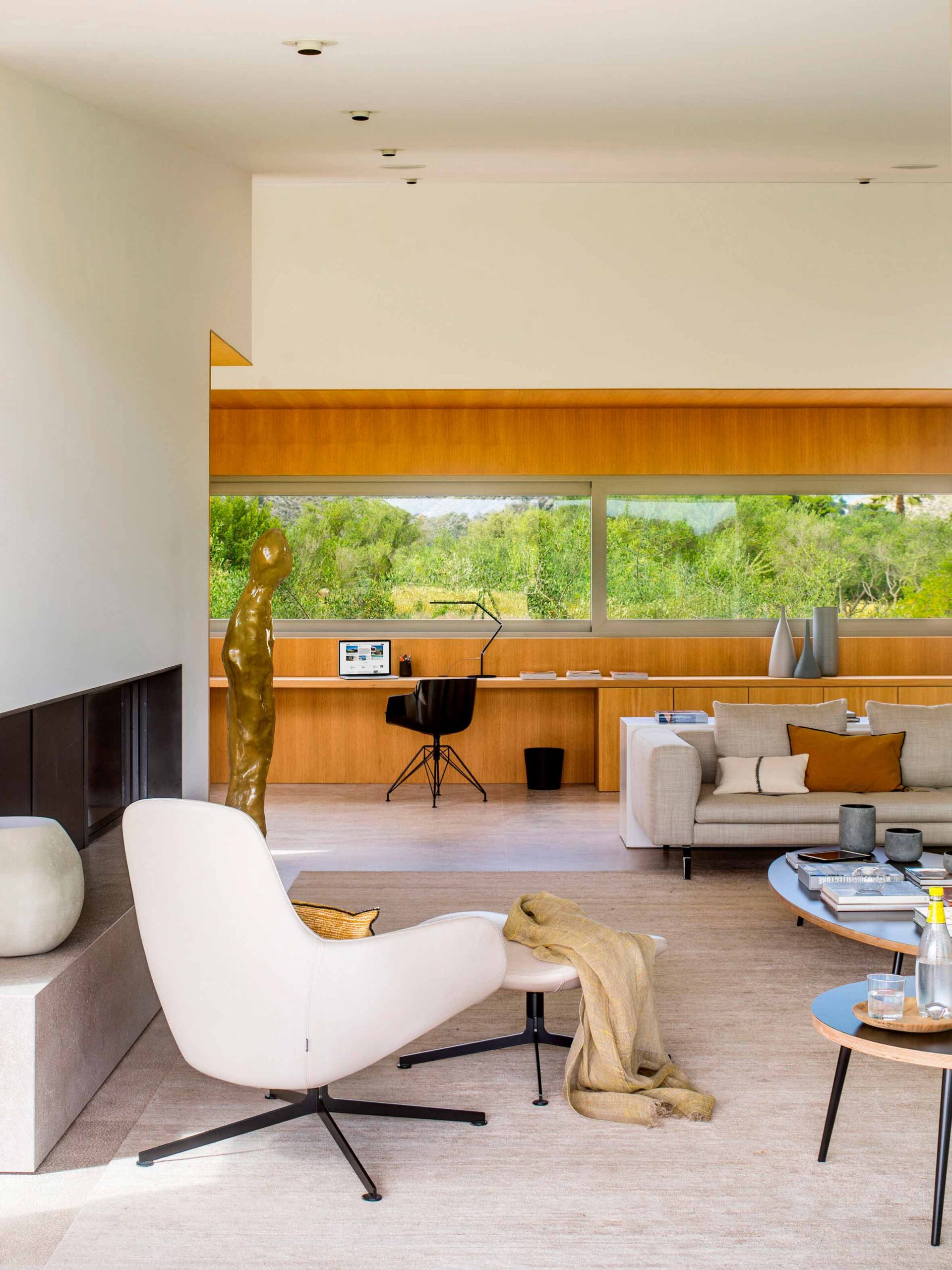 Rincón de despacho en madera clara con ventanal a la vegetación, sofá claro y sillón blanco