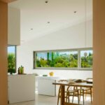 Cocina blanca y comedor vistos entre paneles de madera, con ventana alargada y jardín con palmeras