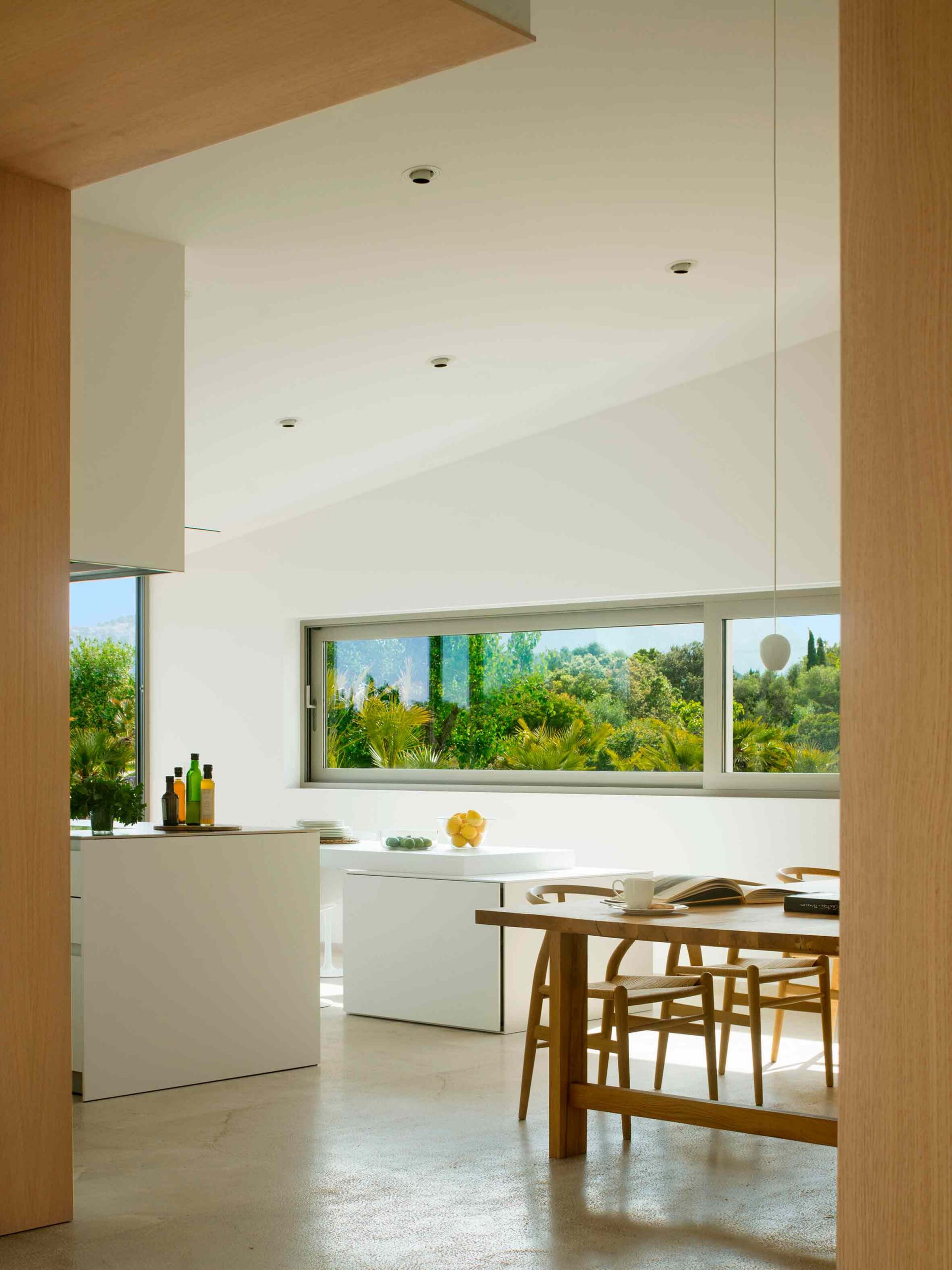 Cocina blanca y comedor vistos entre paneles de madera, con ventana alargada y jardín con palmeras