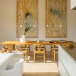 Comedor con mesa de madera y sillas de asiento trenzado bajo dos cuadros dorados, junto a isla blanca