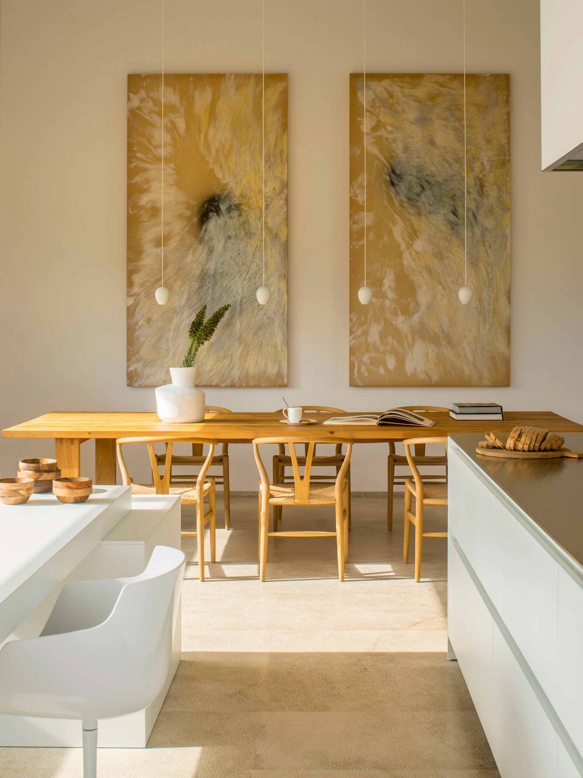 Comedor con mesa de madera y sillas de asiento trenzado bajo dos cuadros dorados, junto a isla blanca