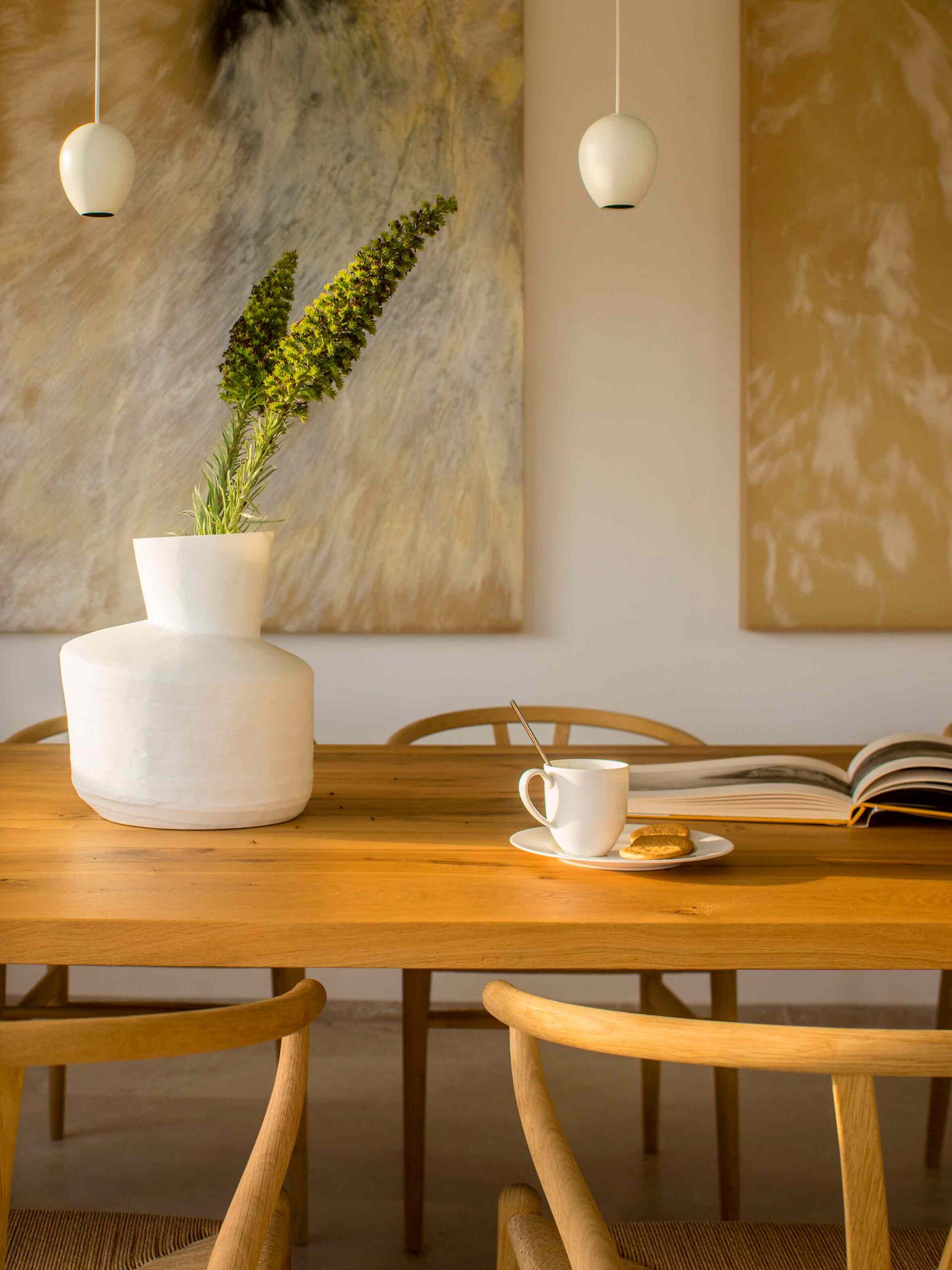 Detalle de la mesa de madera con jarrón blanco, planta verde, taza, libro abierto y lámparas colgantes
