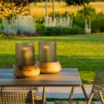 Detalle de dos faroles con velas sobre la mesa exterior, con césped y jardín bajo luz dorada