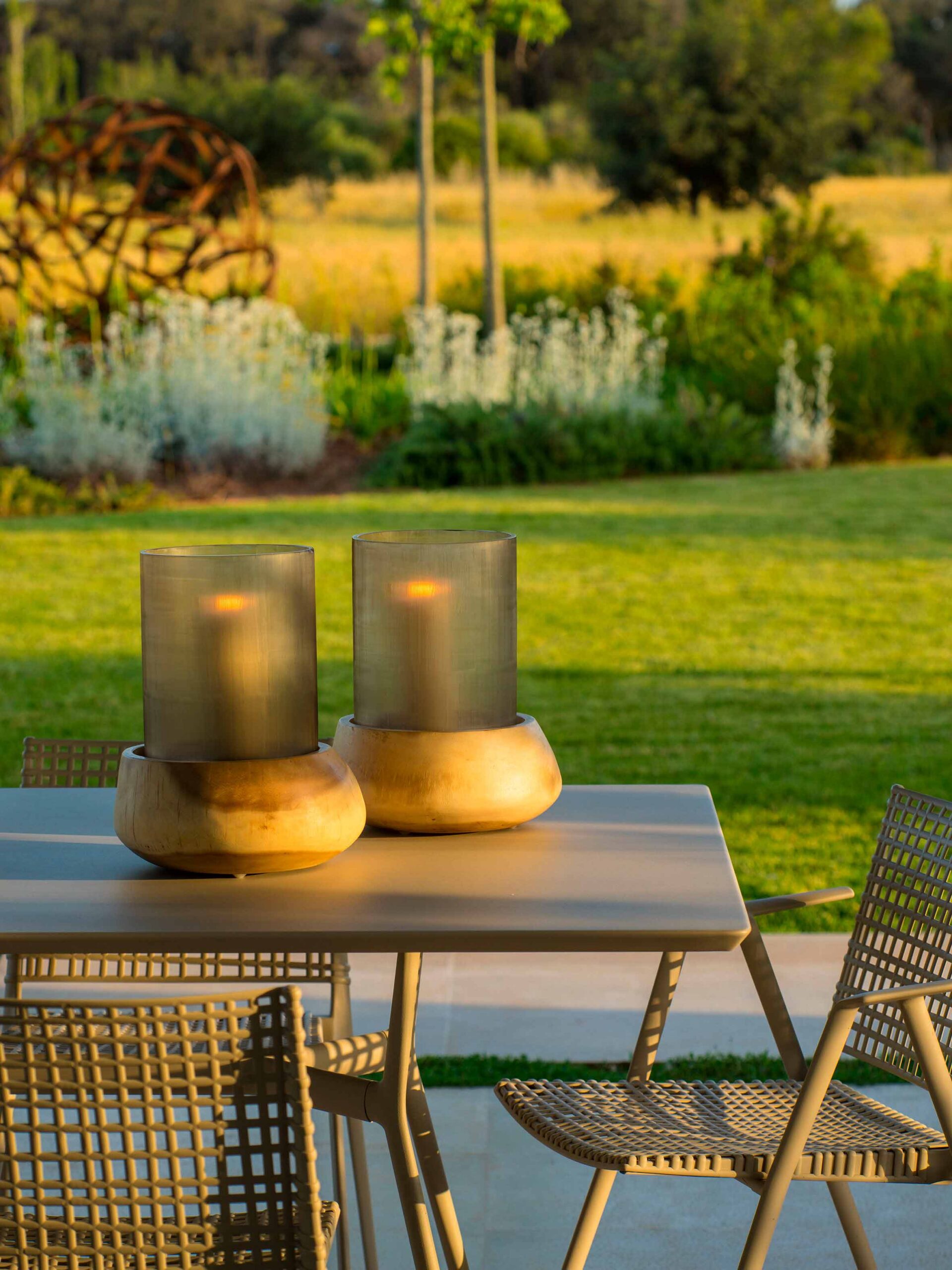 Detalle de dos faroles con velas sobre la mesa exterior, con césped y jardín bajo luz dorada