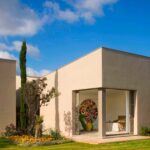 Pabellón del dormitorio con esquina acristalada, fachada de estuco, ciprés, flores amarillas y césped