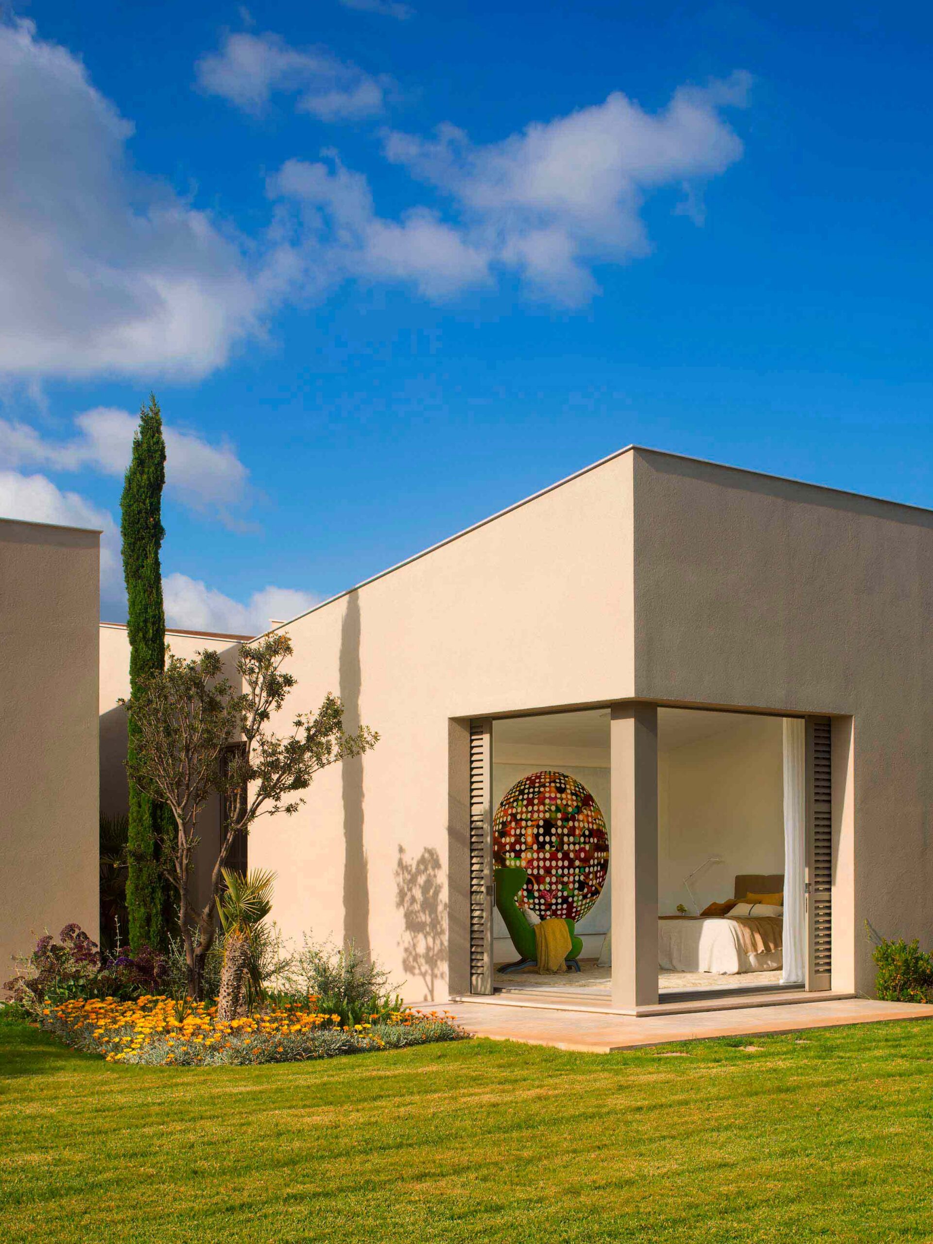 Pabellón del dormitorio con esquina acristalada, fachada de estuco, ciprés, flores amarillas y césped