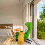 Dormitorio con cama de cabecero tapizado, sillón verde, manta amarilla, cortinas blancas y puerta corredera al jardín