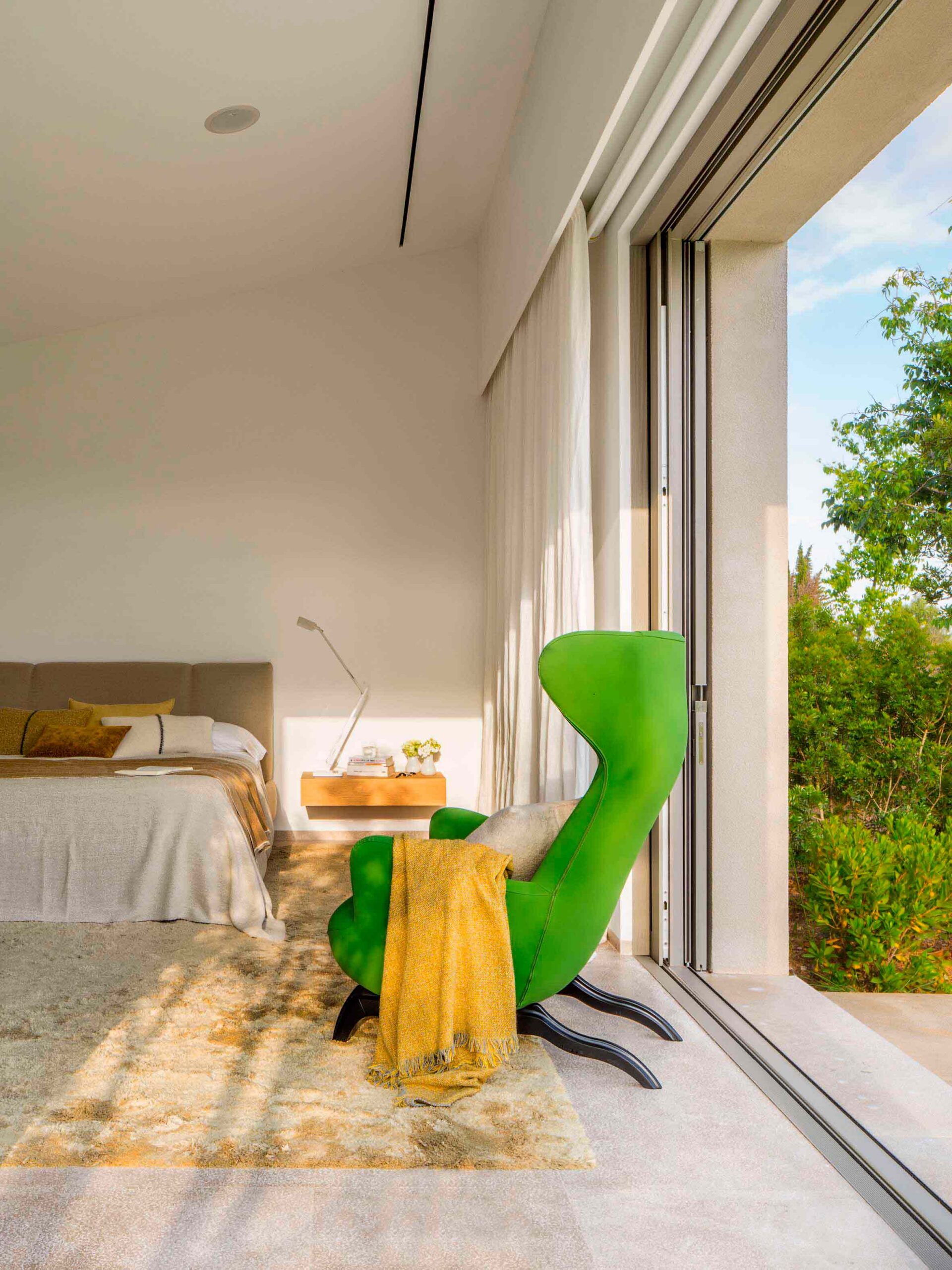 Dormitorio con cama de cabecero tapizado, sillón verde, manta amarilla, cortinas blancas y puerta corredera al jardín