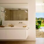 Mueble de baño suspendido blanco con espejo amplio, grifería de pared y sillón verde al fondo