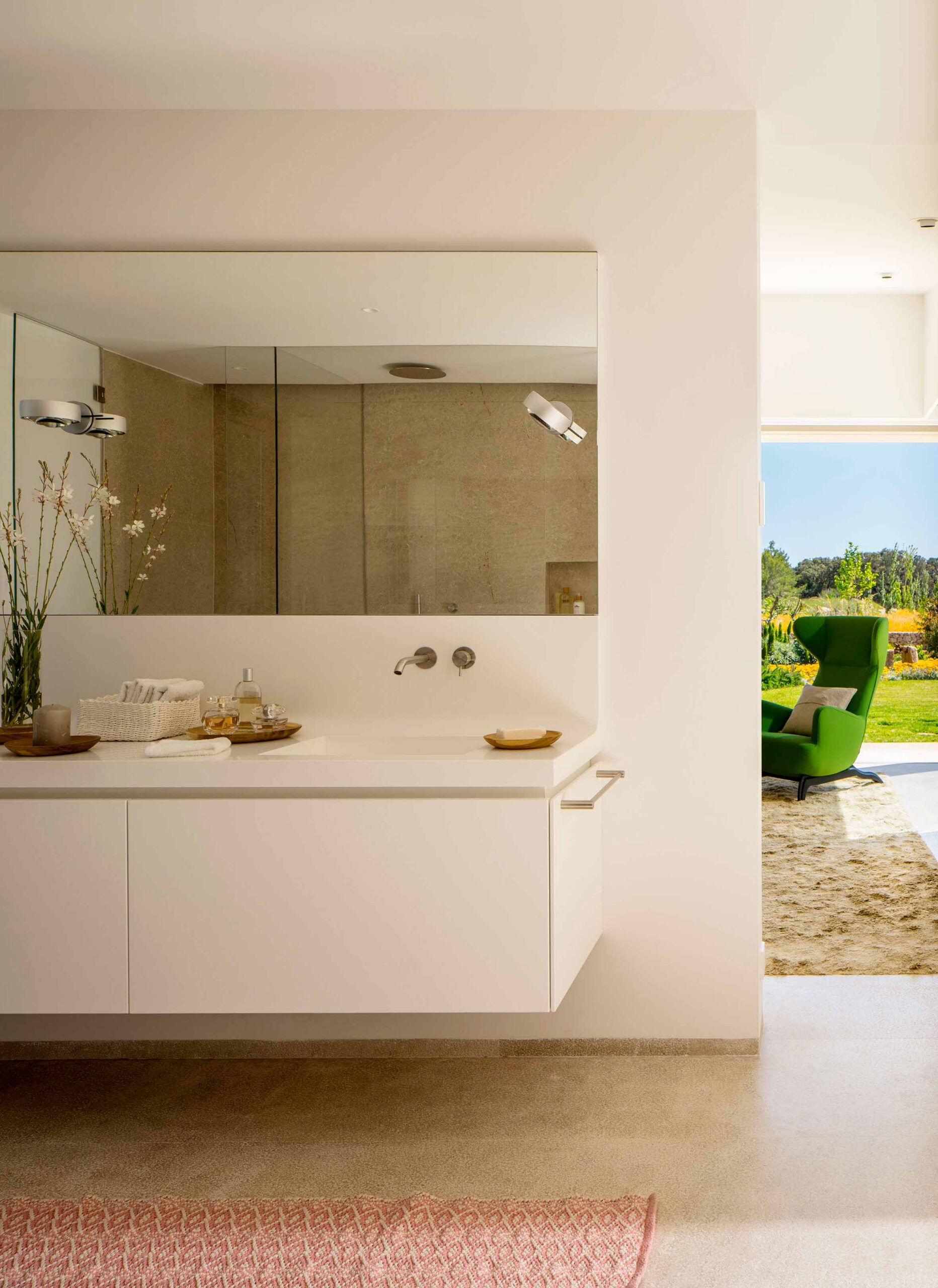 Mueble de baño suspendido blanco con espejo amplio, grifería de pared y sillón verde al fondo