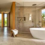 Baño con ducha de cristal revestida de piedra, bañera exenta blanca, taburete de madera y suelo continuo claro
