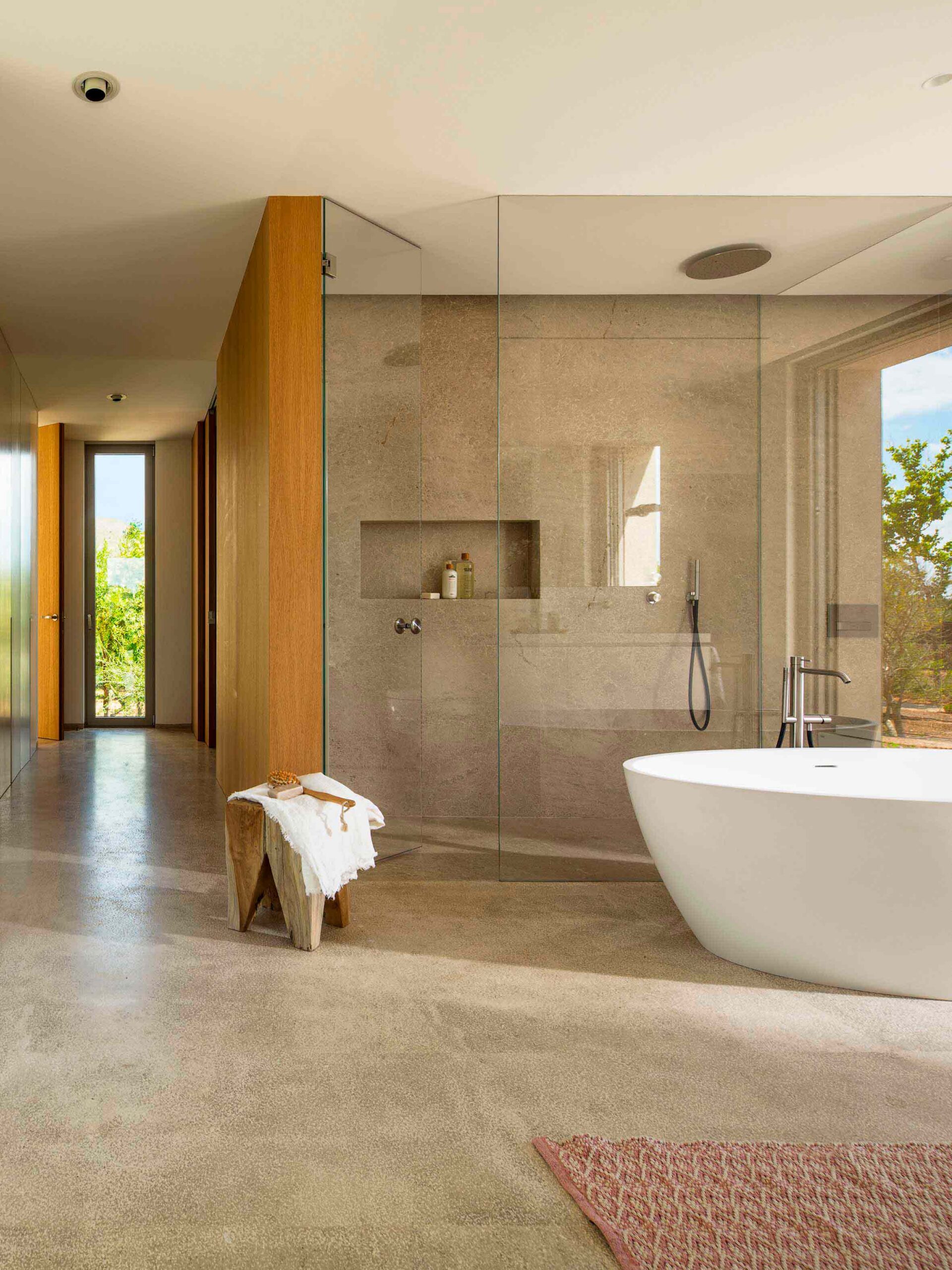 Baño con ducha de cristal revestida de piedra, bañera exenta blanca, taburete de madera y suelo continuo claro