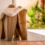 Detalle de taburete de madera natural con toalla blanca y alfombra rosa, con palmeras desenfocadas al fondo