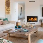 Mesa baja de madera rústica y sofá claro frente a la chimenea, con pasillo al fondo.