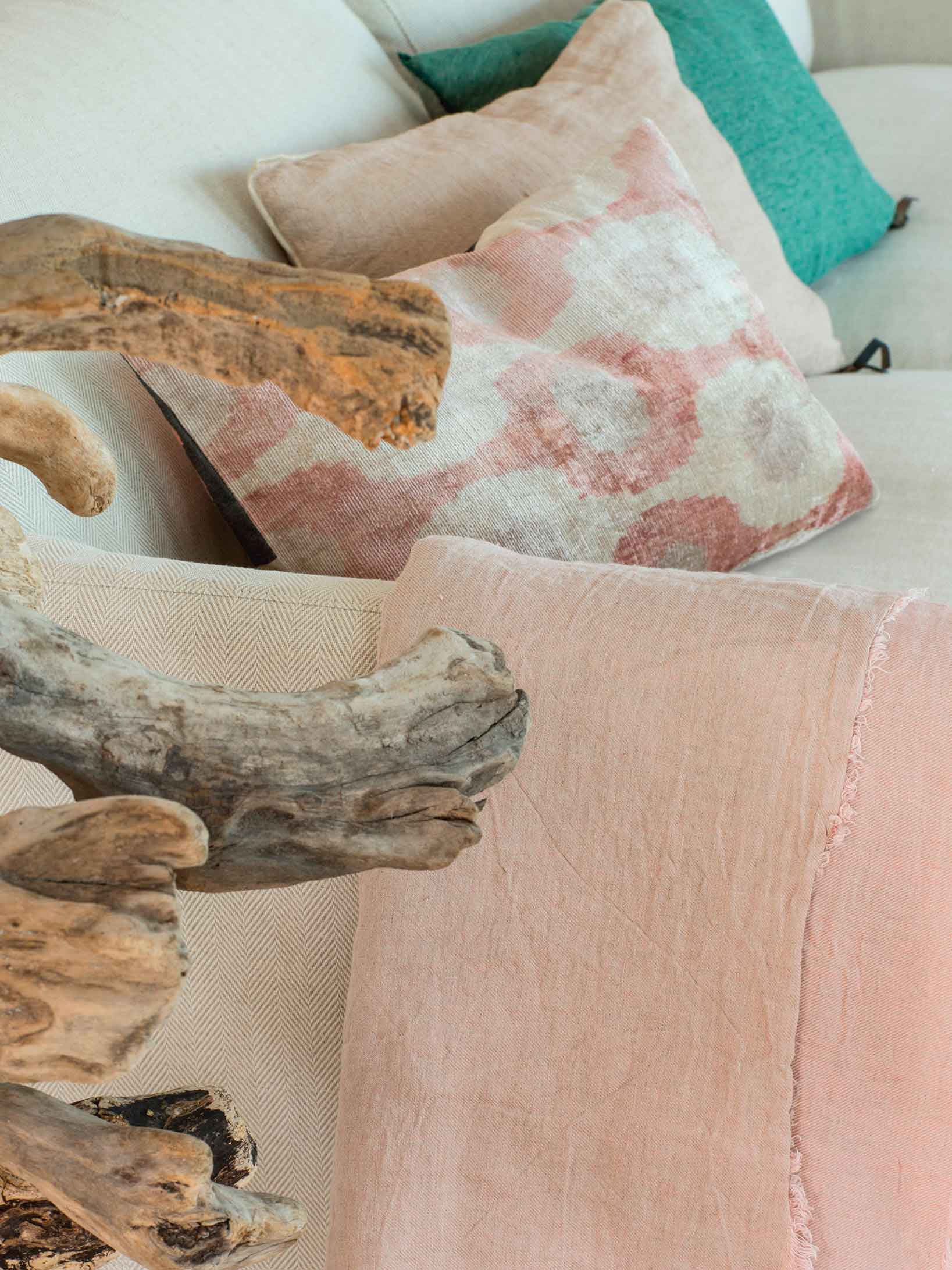 Detalle de cojines rosas y verdes, manta rosada y ramas de madera flotada junto al sofá.