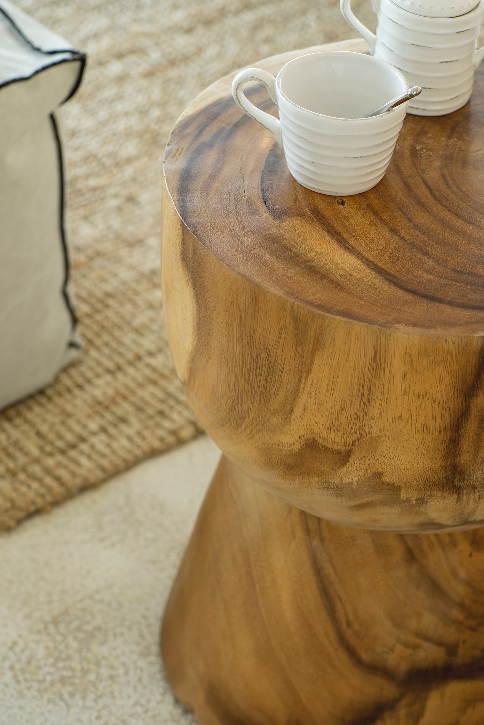 Detalle de mesa auxiliar de madera maciza con taza y tetera blancas, con alfombra de fibra al fondo.