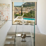 Escalera con barandilla de cristal, lámpara de esferas y gran ventana con vistas a la piscina.