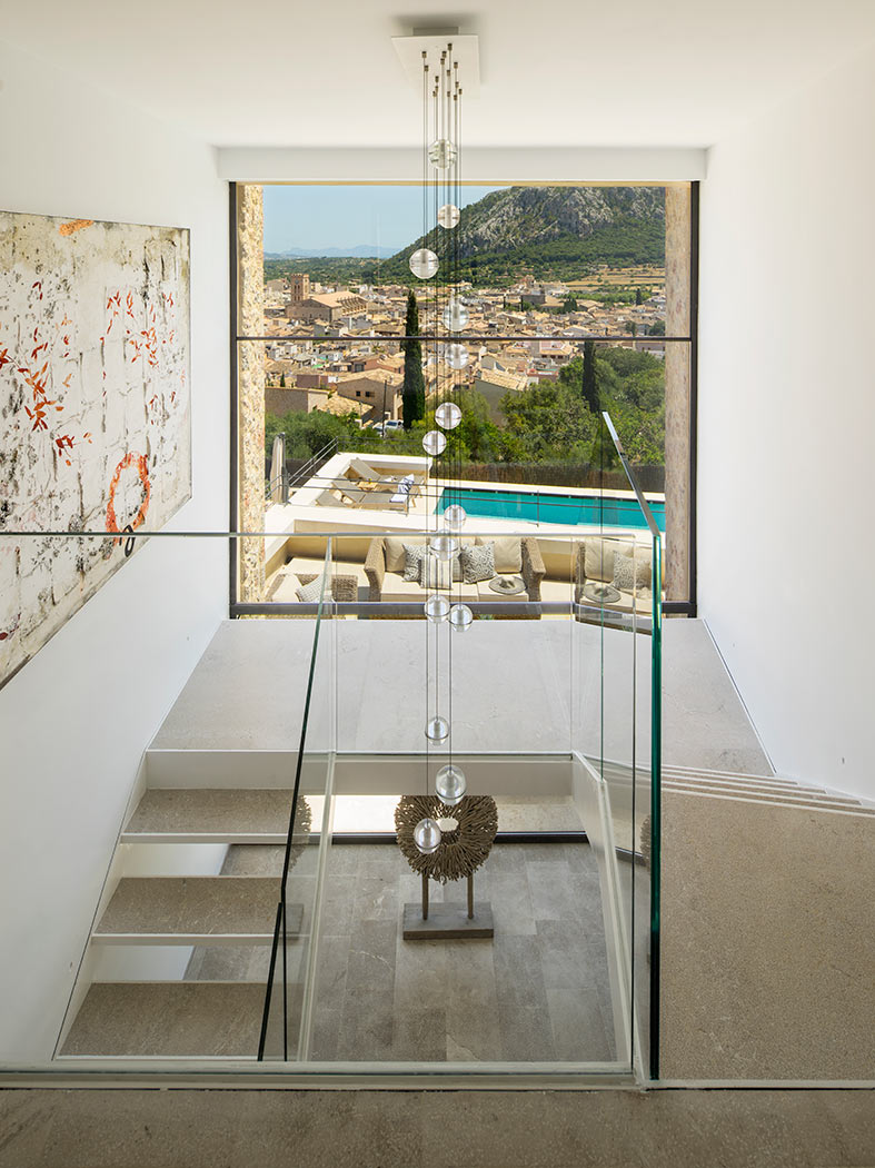 Escalera con barandilla de cristal, lámpara de esferas y gran ventana con vistas a la piscina.