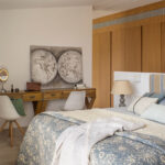 Cama con colcha floral azul junto al escritorio con dos sillas y cuadro de mapamundi.
