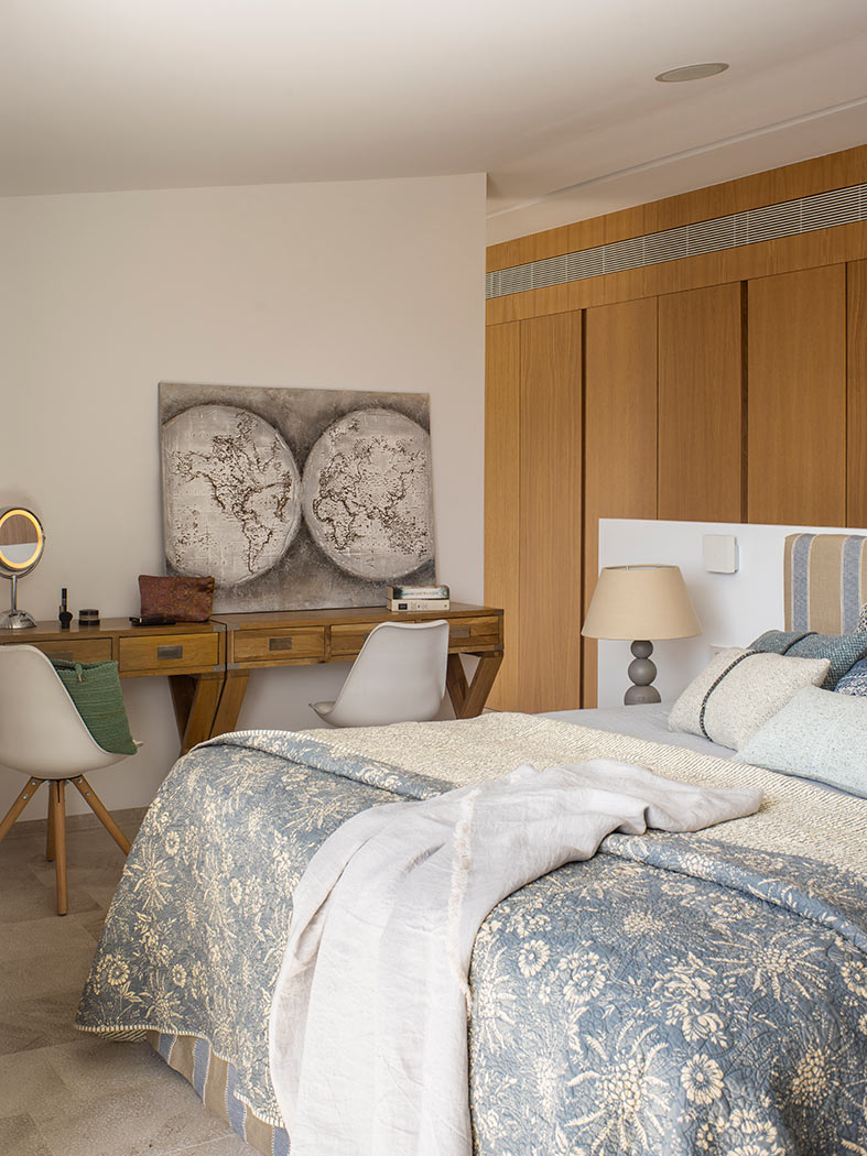 Cama con colcha floral azul junto al escritorio con dos sillas y cuadro de mapamundi.