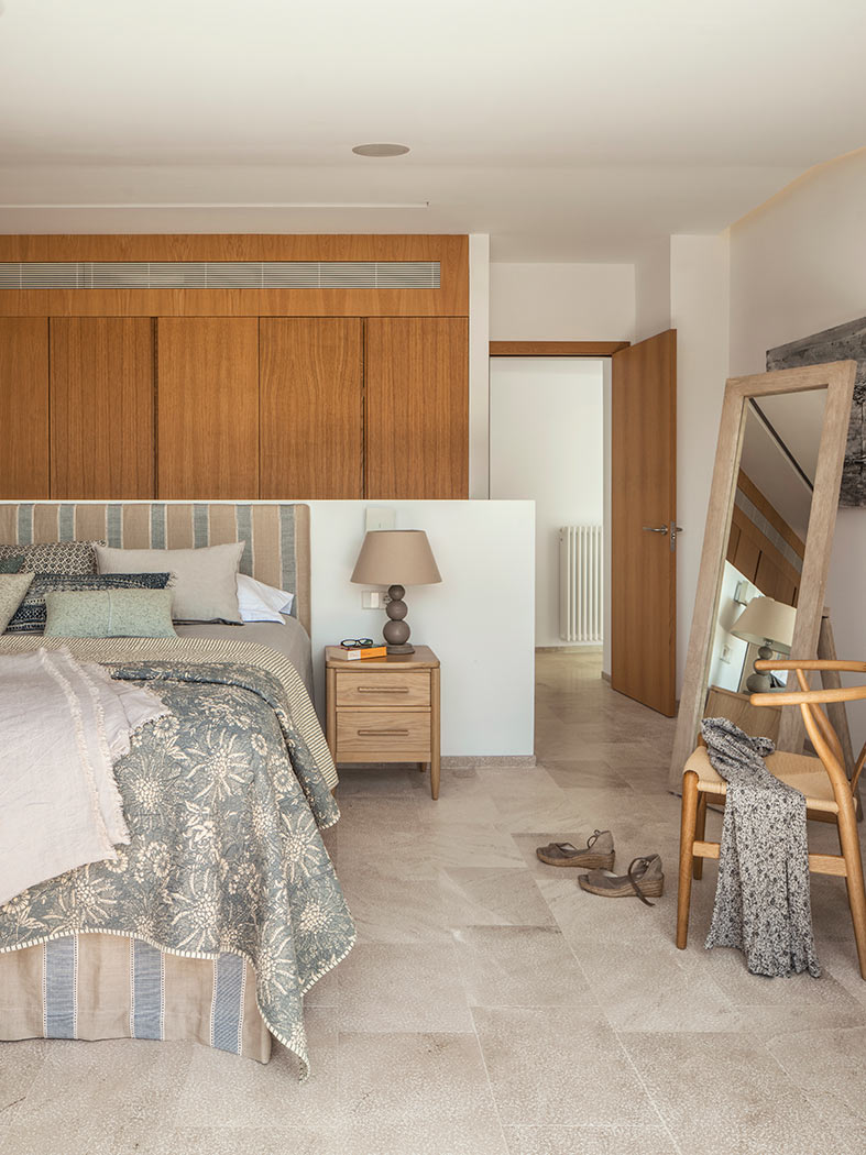 Dormitorio con armarios de madera, mesilla, espejo de pie, silla y suelo de piedra clara.