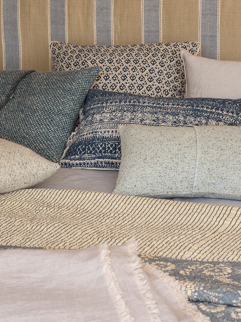 Detalle de cojines estampados en azul y beige, mantas y cabecero de rayas en la cama.