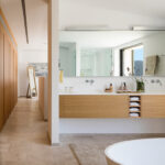 Baño con lavabo doble suspendido de madera clara, espejo grande, bañera blanca y suelo de piedra.