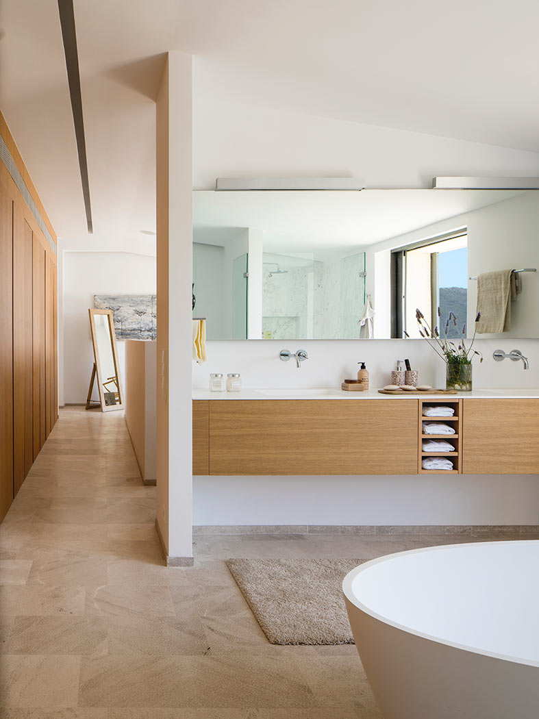Baño con lavabo doble suspendido de madera clara, espejo grande, bañera blanca y suelo de piedra.