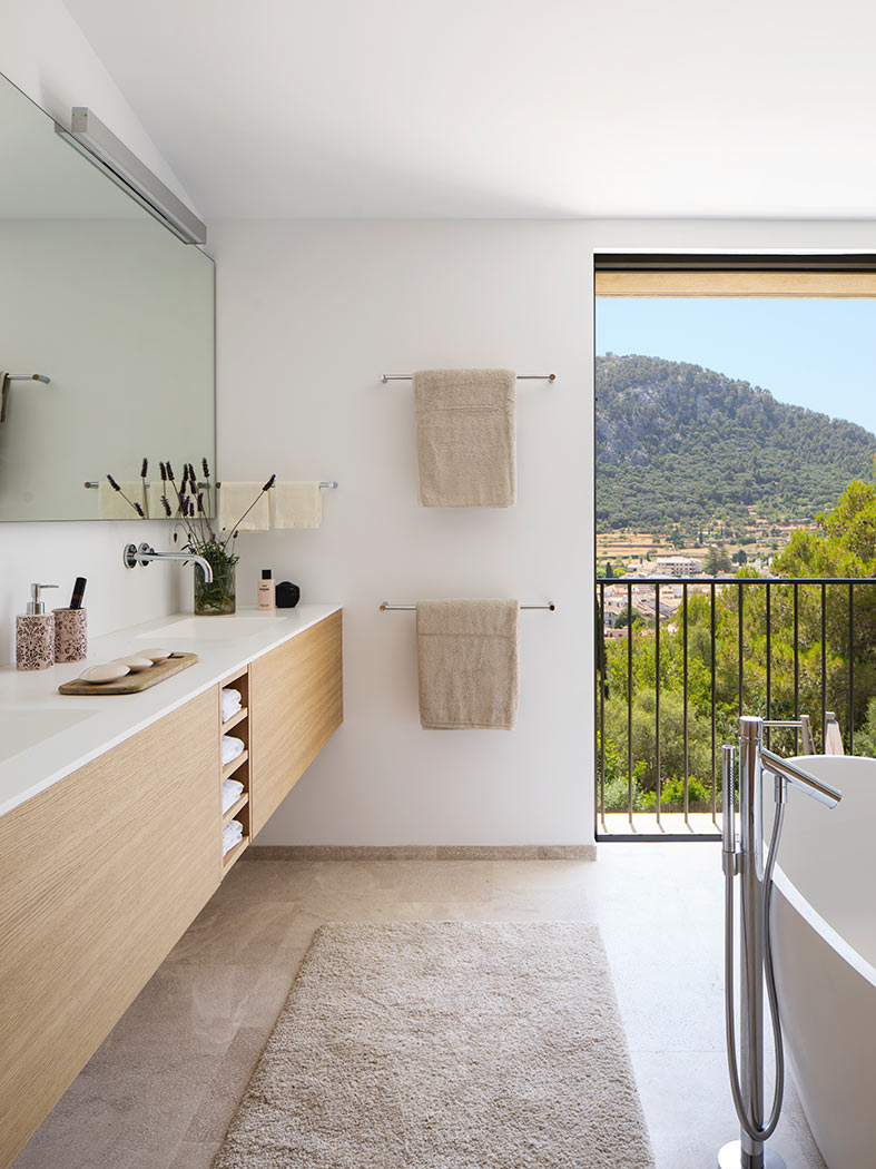 Mueble de lavabo de madera clara, toallas y ventanal con balcón y vistas a la montaña.