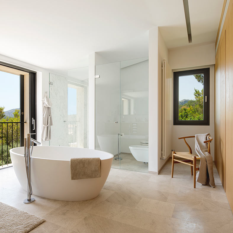 Baño amplio con bañera blanca junto al balcón, silla de madera y ducha de cristal al fondo.