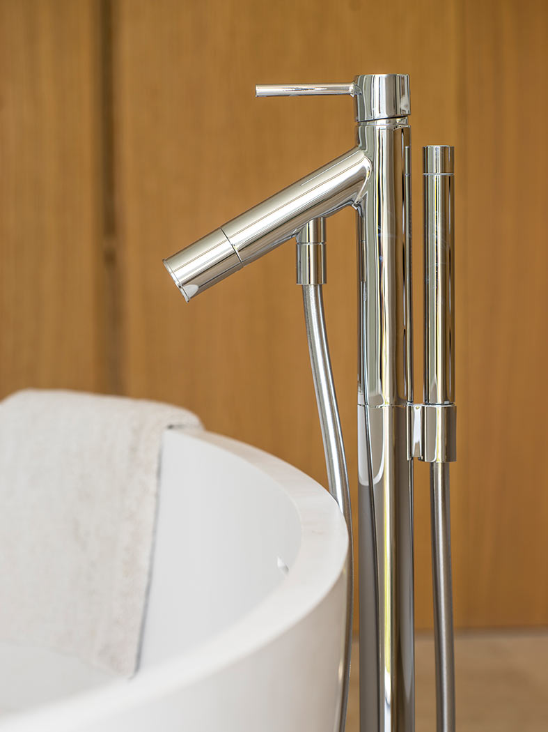 Detalle del grifo cromado de bañera exenta con ducha de mano sobre fondo de madera clara.