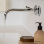 Detalle de grifo cromado de pared en el lavabo, con agua corriendo y productos de baño.