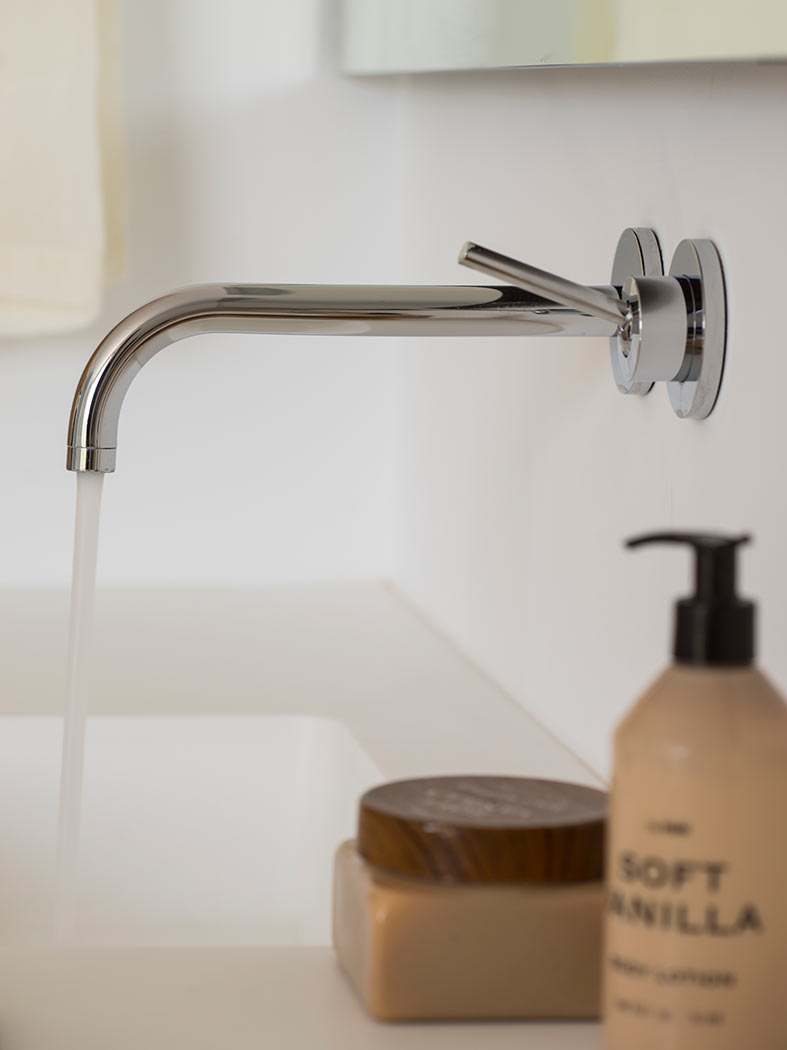 Detalle de grifo cromado de pared en el lavabo, con agua corriendo y productos de baño.