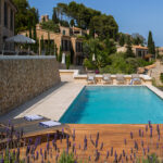 Piscina con tarima de madera, lavanda en primer plano, tumbonas, muro de piedra y casas al fondo.
