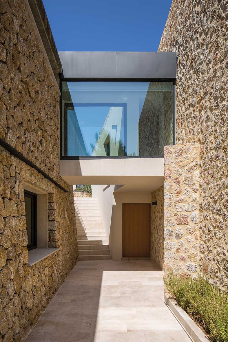 Pasaje entre muros de piedra con pasarela acristalada, puerta de madera, escalera y suelo de piedra clara.