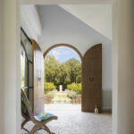 Entrada con puertas de madera en arco abiertas al jardín, suelo de guijarros y sillón de tapizado floral