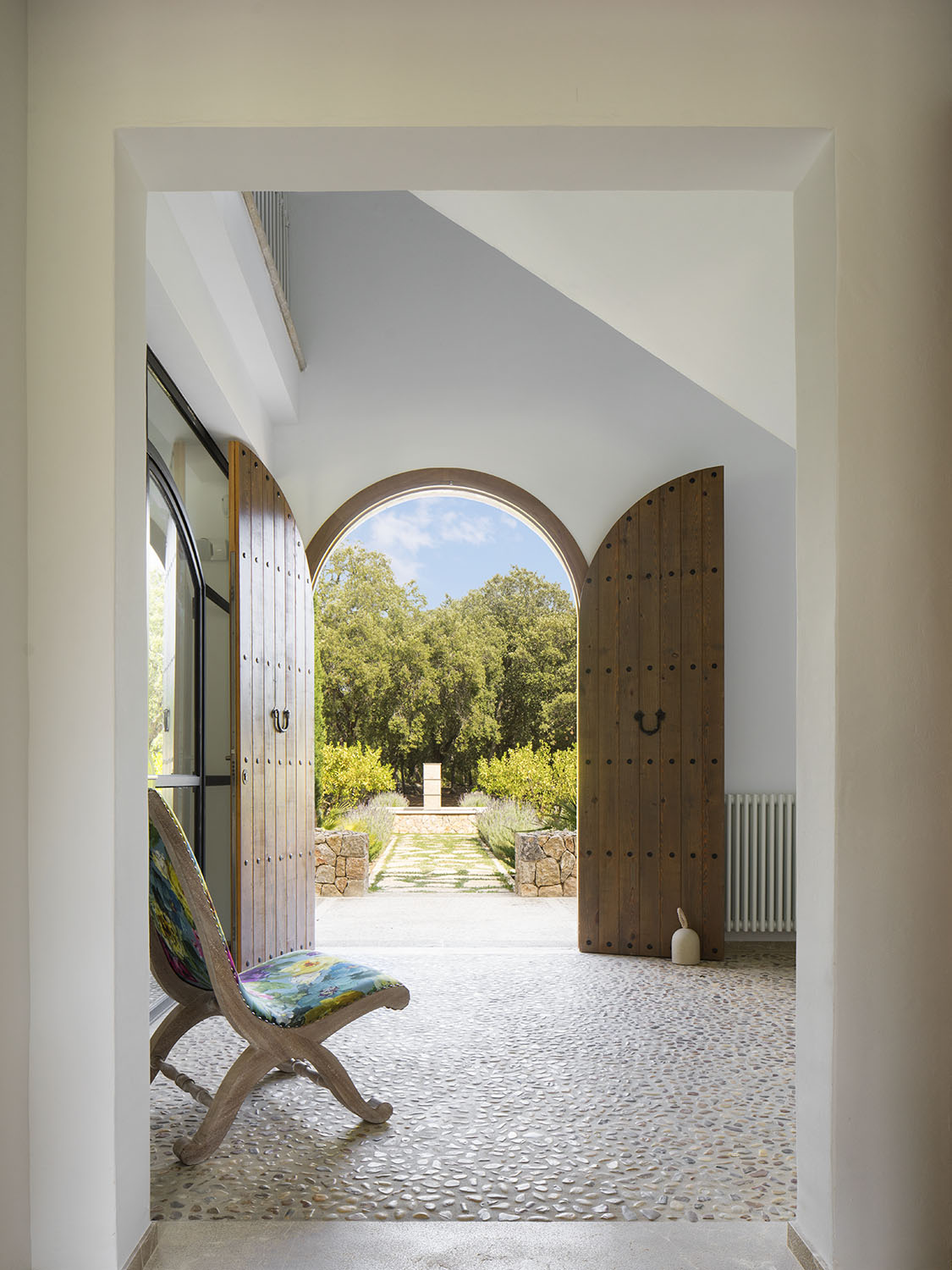 Entrada con puertas de madera en arco abiertas al jardín, suelo de guijarros y sillón de tapizado floral