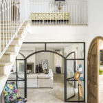 Vestíbulo con escalera de barandilla blanca, cristalera de hierro con arcos, suelo de guijarros y sillón floral